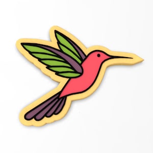 May include: A yellow cookie decorated with a pink hummingbird with green wings. The hummingbird is flying with its wings spread out.