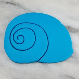 May include: A bright turquoise, shell-shaped craft element with a spiral design. The shell is a solid color with a darker blue outline, and it sits on a white and gray marble surface. This item is likely used for crafting or decoration.