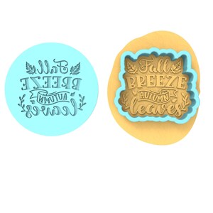 Op de afbeelding: Een blauwe koekjesvormer met de tekst "Fall Breeze Autumn Leaves" in een decoratief lettertype. De vormer ligt op een bruin oppervlak.