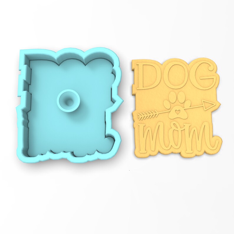 May include: A blue plastic cookie cutter shaped like the word "Dog Mom" with an arrow and paw print design. The cutter is next to a yellow cookie with the same design.
