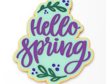 May include: A light blue cookie decorated with purple icing and the words "Hello Spring" in a cursive font. The cookie is also decorated with green leaves and purple dots.