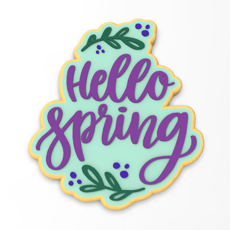 Op de afbeelding: Een lichtblauwe koek versierd met paarse glazuur en de woorden "Hello Spring" in een cursief schrift. De koek is ook versierd met groene bladeren en paarse stippen.