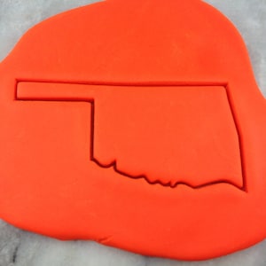 May include: A red cookie cutter in the shape of the state of Oklahoma. The cutter is on a red surface.
