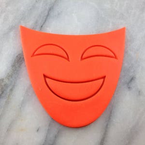 May include: A bright orange plastic cookie cutter shaped like a smiling face with a wide mouth and closed eyes.