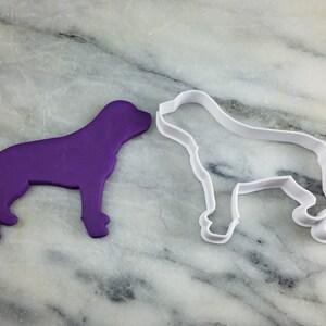 K&ouml;nnte beinhalten: Ein violetter Plastik-Keksstempel in Form eines Hundes und ein wei&szlig;er Plastik-Keksstempel in Form eines Hundes auf einer Marmorfl&auml;che.