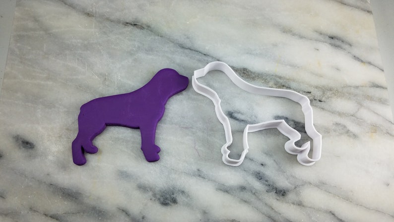 K&ouml;nnte beinhalten: Ein violetter Plastik-Keksstempel in Form eines Hundes und ein wei&szlig;er Plastik-Keksstempel in Form eines Hundes auf einer Marmorfl&auml;che.