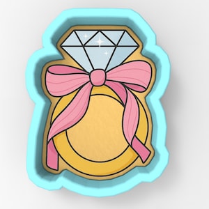 May include: A cookie cutter in the shape of a diamond ring with a pink bow. The ring is gold with a large diamond on top. The cutter is surrounded by a light blue border.