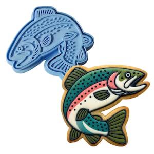 May include: A blue plastic fish-shaped cookie cutter next to a decorated cookie. The cookie is shaped like a fish with green, red, and white details. The cookie has a brown outline and is decorated with a rainbow pattern.