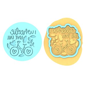 Op de afbeelding: Een blauwe koekjesvormer met de tekst "I wheelie love fall" en een fiets met bladeren en harten.