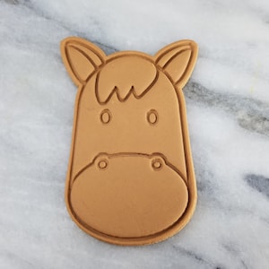 Puede incluir: Un cortador de galletas de plástico marrón con forma de cabeza de caballo. El cortador tiene un diseño simple con orejas redondeadas, ojos y nariz.