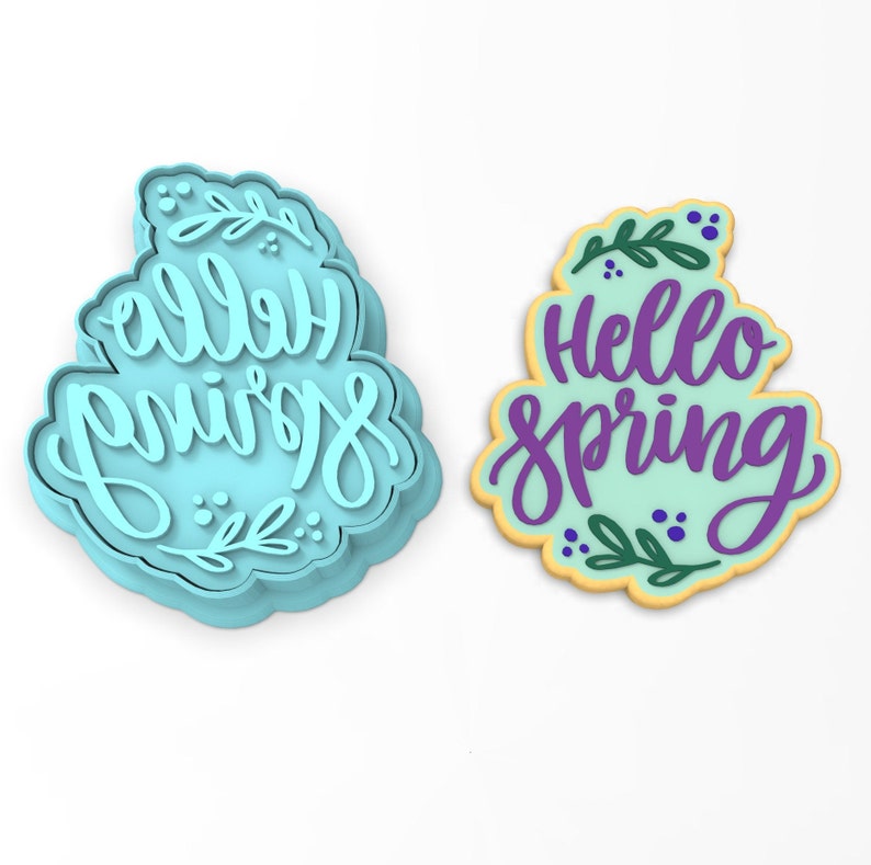 Op de afbeelding: Twee koekjesvormen, een blauwe en een gele, met de tekst "hello spring" in paarse letters. De vormen zijn versierd met groene bladeren en kleine blauwe stippen.