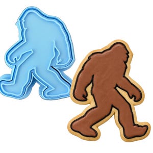 May include: A light blue plastic cookie cutter shaped like a walking Bigfoot silhouette next to a matching cookie decorated with brown icing and a black piped outline, both depicting the same creature in a side profile walking stance.