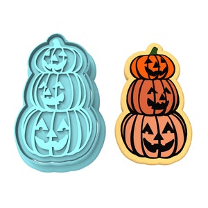 Op de afbeelding: Een blauwe koekjesvormer in de vorm van een stapel van drie jack-o'-lantern pompoenen. De vormer wordt getoond naast een gedecoreerde koek met hetzelfde ontwerp.
