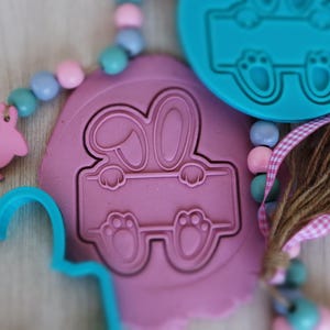 Peut inclure: Deux moules en silicone pour la cuisson, un rose et un bleu. Le moule rose est en forme de lapin qui regarde par-dessus un panneau. Le moule bleu est en forme de lapin avec ses pattes qui dépassent.
