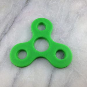 May include: A vibrant green fidget spinner, featuring three curved arms and circular openings. The toy is made of a solid material and is set against a marble background.