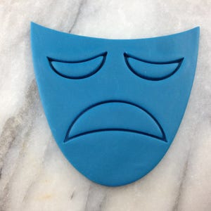 May include: A blue, teardrop-shaped mask with a sad face design. The mask features a curved mouth and two curved lines for eyes. The mask is a solid, matte blue color.