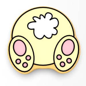 May include: A yellow bunny cookie with pink ears and a white fluffy tail. The cookie has a black outline and is decorated with three black paw prints on each foot.