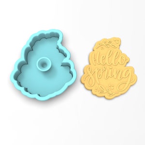 Op de afbeelding: Een lichtblauwe plastic koekjesvormer in de vorm van een wolk met de tekst "Hello Spring" erin. Naast de vormer is een gele koek te zien, met hetzelfde ontwerp.