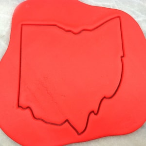May include: A red cookie cutter in the shape of the state of Ohio. The cutter is on a red surface.