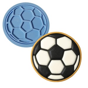 May include: A blue silicone mold and a decorated cookie in the shape of a soccer ball. The cookie has a black and white pentagon pattern with a yellow border. The mold is round with a soccer ball design.