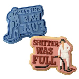 May include: A blue cookie cutter and a decorated cookie. The cookie cutter is blue and shaped like a person with the text "SHITTER WAS FULL" in reverse. The cookie is decorated with the same text and a person.