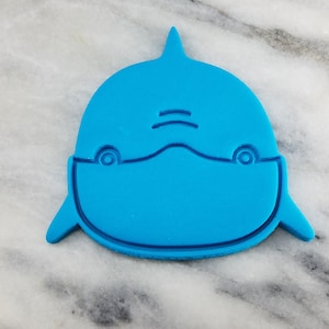 Op de afbeelding: Een blauwe plastic koekjesvormer in de vorm van een dolfijn met een eenvoudig, cartoonachtig gezicht.