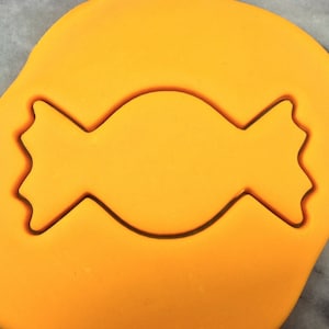 May include: A yellow fondant cookie cutter in the shape of a candy. The cutter is on a yellow fondant surface.