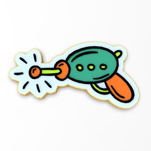 May include: A cartoon-style toy gun cookie cutter with a green body, orange handle, and yellow details. The cutter is shooting a beam of light.