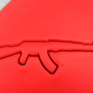 以下が含まれることがあります： 赤色の銃の形をしたクッキーカッター。カッターはAK-47アサルトライフルのシルエットです。デザインは赤い素材に彫り込まれ、銃器のネガティブスペースの輪郭を作成しています。