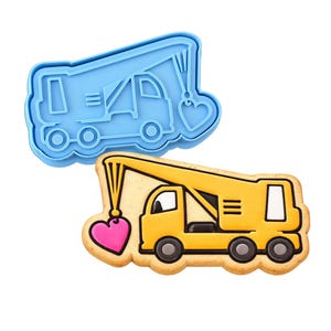 May include: A yellow crane truck cookie with a pink heart-shaped pendant, next to a blue plastic cookie cutter in the shape of the crane truck. The cookie has a light brown border and black wheels. The crane arm is extended.