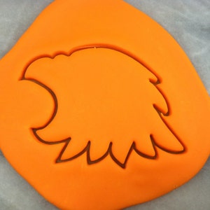May include: Orange fondant with an eagle cookie cutter pressed into it. The cutter is shaped like an eagle's head with a beak and feathers.