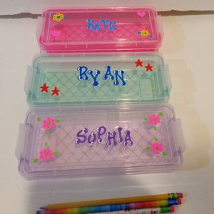 May include: Three clear plastic pencil cases with personalised names. The top case is pink with the name "KATE" in blue letters. The middle case is light green with the name "RYAN" in blue letters. The bottom case is lavender with the name "SOPHIA" in purple letters. Each case has a decorative design.