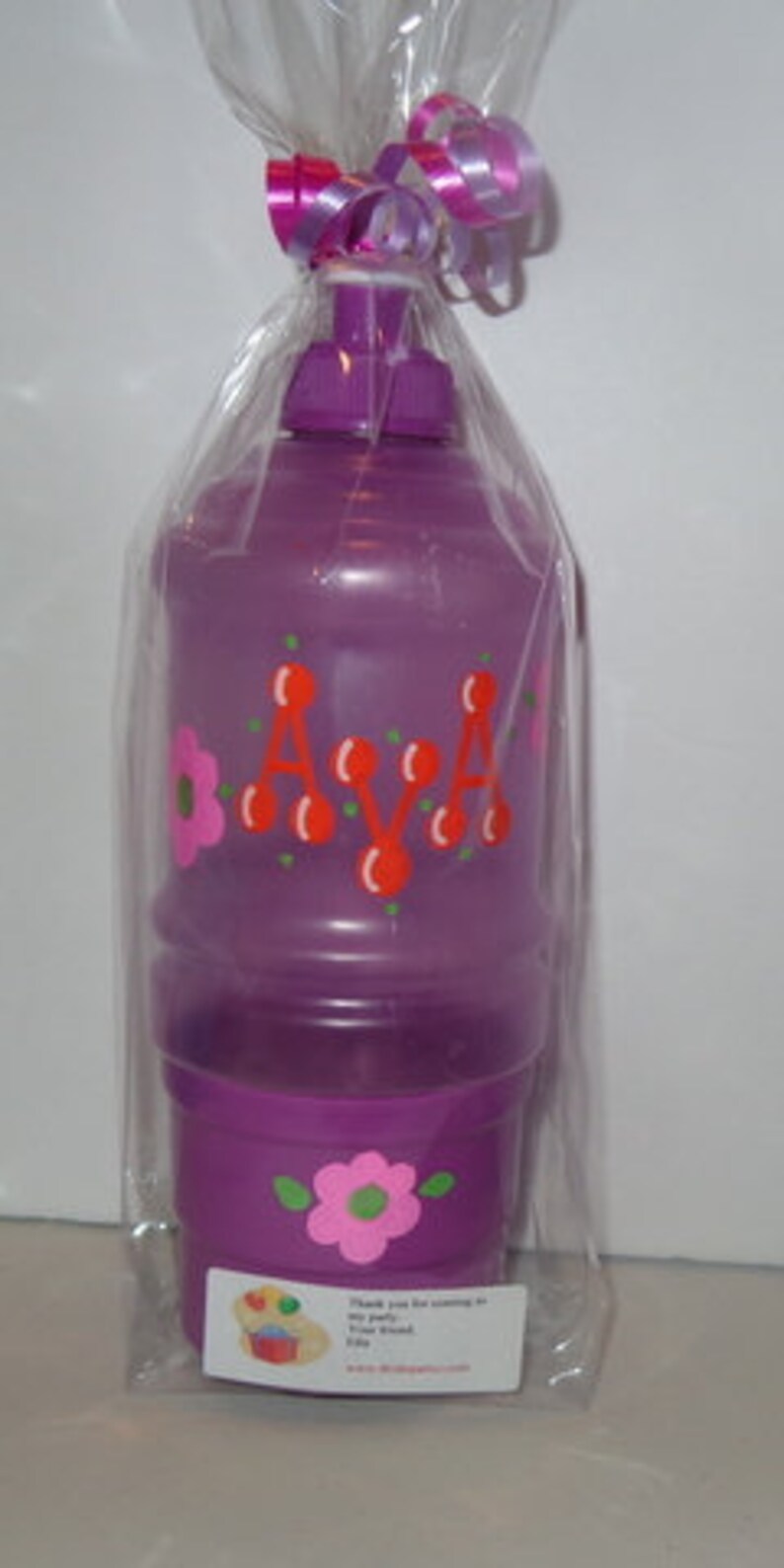 May include: A purple water bottle with a lid and a cup attached. The bottle is decorated with red cherries and the name "AVA" in orange letters. The cup has a pink flower design.