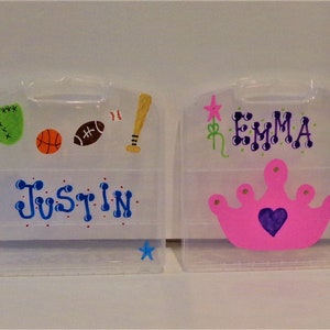 Op de afbeelding: Twee doorzichtige plastic containers met gepersonaliseerde ontwerpen. De container aan de linkerkant heeft een honkbal, een basketbal, een voetbal en een honkbalknuppel erop geschilderd. De container aan de rechterkant heeft een roze kroon met een paars hart en de naam "EMMA" erop geschilderd.