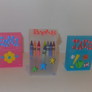 May include: Three personalized crayon boxes, one pink with a flower design and the name "Emma" in blue, one clear with stars and the name "Ryan" in red, and one blue with sports designs and the name "Jake" in red.