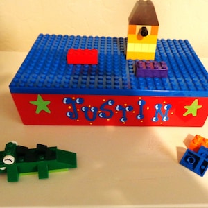 May include: A red, blue, and yellow Lego toy box with the name "Justin" painted on the front. The box is decorated with stars and has a blue Lego base with a red, blue, and green Lego piece on top.