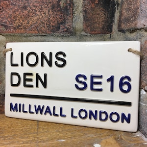 Peut inclure: Panneau rectangulaire en céramique blanche avec l'inscription "LIONS DEN SE16 MILLWALL LONDON" en lettres noires et bleues. Le panneau est suspendu par une ficelle. Le fond est un mur de briques.