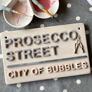 May include: A rectangular ceramic sign with the words "PROSECCO STREET CITY OF BUBBLES" in raised gray and pink letters. Two champagne flutes are depicted. Paintbrushes and a paint palette with pink paint are in the background.