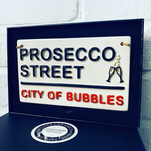 May include: A ceramic street sign with the words "PROSECCO STREET CITY OF BUBBLES" in black and red lettering. The sign includes two champagne glasses and is framed in black.