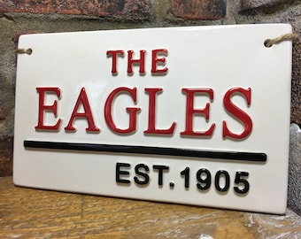 May include: White rectangular sign with black and red lettering. The sign reads "The Eagles Est. 1905".