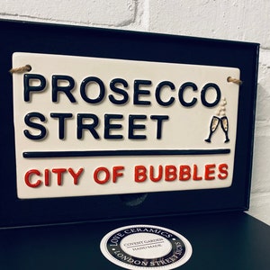 May include: A ceramic street sign that reads "PROSECCO STREET CITY OF BUBBLES" in black and red lettering. The sign has a cream background and a black border. Two champagne glasses are depicted. The sign is in a black box.
