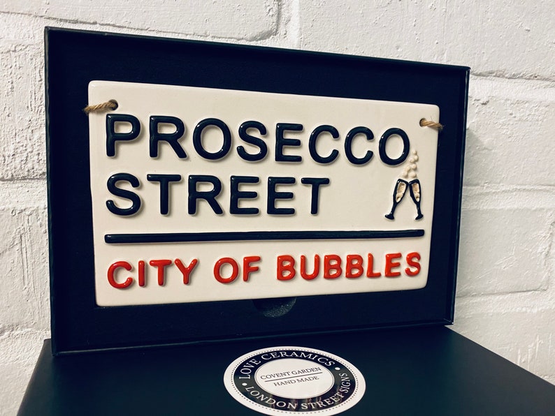 May include: A ceramic street sign that reads "PROSECCO STREET CITY OF BUBBLES" in black and red lettering. The sign has a cream background and a black border. Two champagne glasses are depicted. The sign is in a black box.