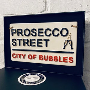 May include: A ceramic street sign in a black frame reads "PROSECCO STREET CITY OF BUBBLES" in black and red lettering. Two champagne glasses are depicted. The sign is cream-colored with a rope for hanging.