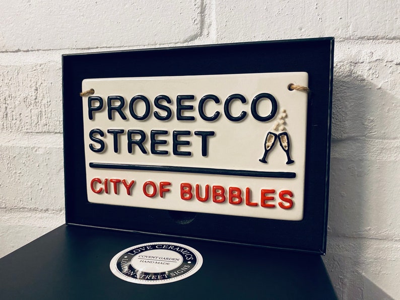 May include: A ceramic street sign in a black frame reads "PROSECCO STREET CITY OF BUBBLES" in black and red lettering. Two champagne glasses are depicted. The sign is cream-colored with a rope for hanging.