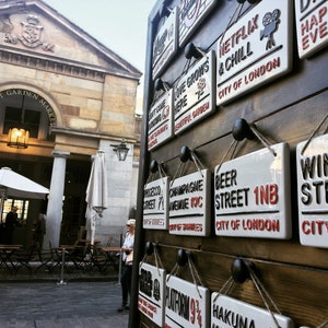 May include: A display of ceramic street signs with various phrases and locations, including "Netflix & Chill" and "Beer Street 1NB, City of London." The signs are white with black and red lettering, mounted on a wooden surface.