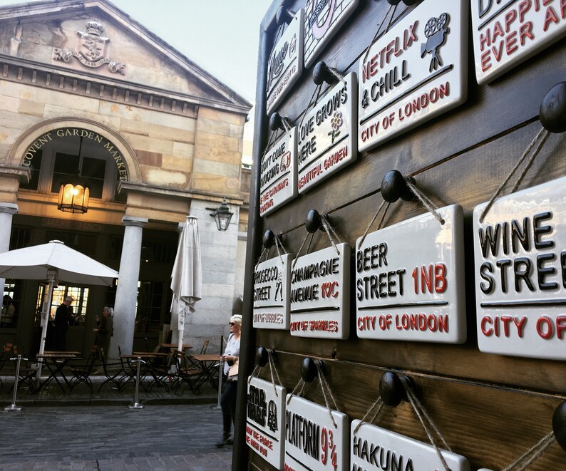 May include: A display of ceramic street signs with various phrases and locations, including "Netflix & Chill" and "Beer Street 1NB, City of London." The signs are white with black and red lettering, mounted on a wooden surface.