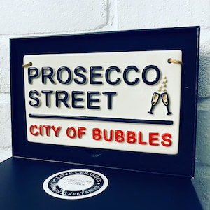 May include: A ceramic street sign with the words "PROSECCO STREET CITY OF BUBBLES" in black and red lettering. The sign includes two champagne glasses and is framed in black.