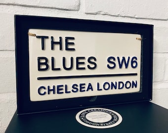 Puede incluir: Un letrero de cerámica blanca con letras negras y azules que dice "The Blues SW6 Chelsea London". El letrero está enmarcado en un marco negro y tiene una pequeña pegatina redonda en blanco y negro que dice "Love Ceramics Covent Garden Handmade London Street Signs".