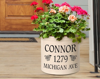 May include: A white fabric planter with black text that reads "CONNOR 1279 MICHIGAN AVE". The planter is filled with pink and red flowers and green foliage. The planter is sitting on a wooden porch.