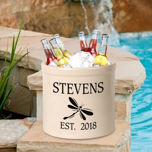 Puede incluir: Una nevera de cerámica blanca con un diseño de libélula negra y el texto "Stevens Est. 2018". La nevera está llena de hielo y botellas de cerveza.